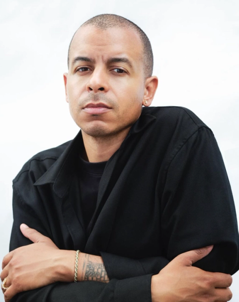A man with a shaved head and light brown skin, wearing a black shirt, crosses his arms and looks seriously at the camera. He wears a gold earring and a gold bracelet.