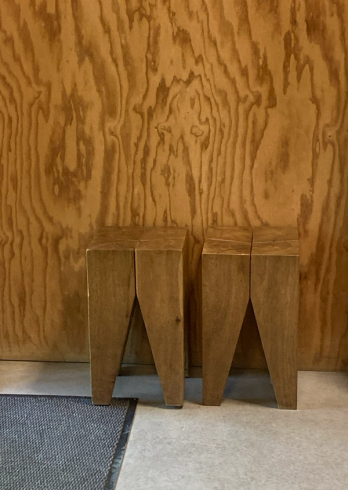 Two wooden stools with V-cut legs against a plywood wall.