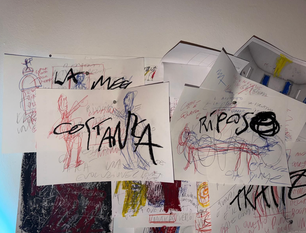 A wall covered with overlapping papers featuring handwritten words like "COSTANZA" and "RIPOSO," along with colorful scribbled drawings.