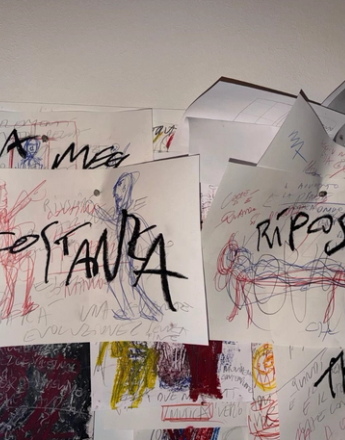 A wall covered with overlapping papers featuring handwritten words like "COSTANZA" and "RIPOSO," along with colorful scribbled drawings.