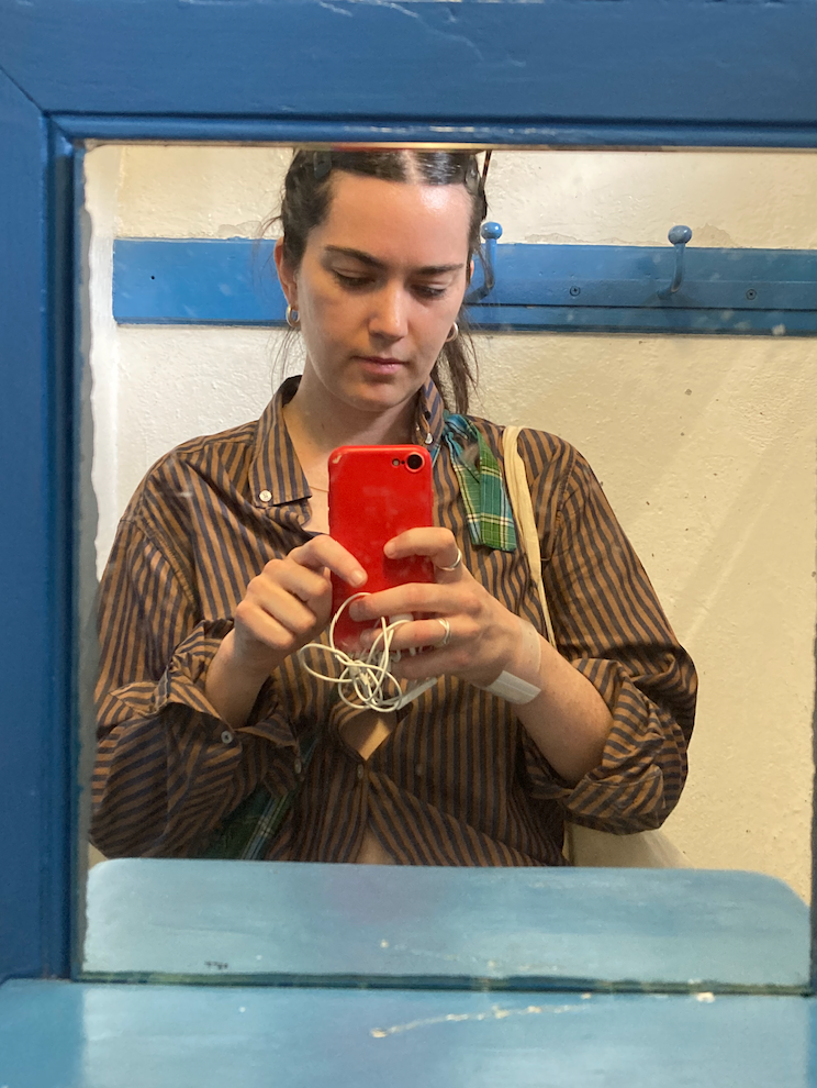 A woman in a striped shirt takes a mirror selfie with a red phone, a bandage visible on her wrist, reflected in a blue-framed mirror.