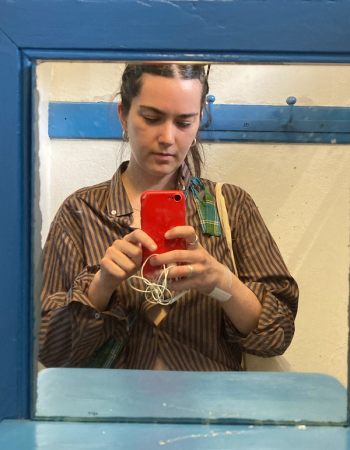 A woman in a striped shirt takes a mirror selfie with a red phone, a bandage visible on her wrist, reflected in a blue-framed mirror.
