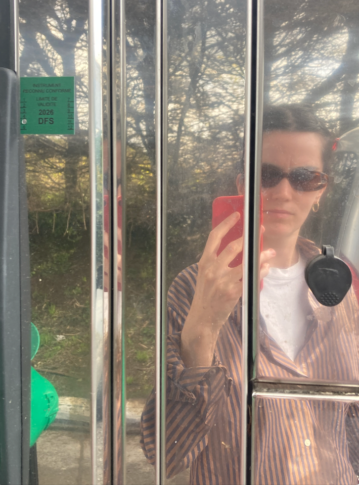 A person in sunglasses and a striped shirt holds a red phone, reflected in a shiny metallic surface.
