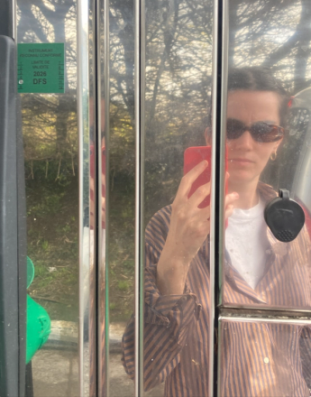 A person in sunglasses and a striped shirt holds a red phone, reflected in a shiny metallic surface.
