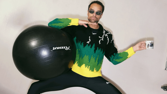 A man in sunglasses and a black, green, and yellow knitted sweater poses dynamically with a large black exercise ball and a white mug.