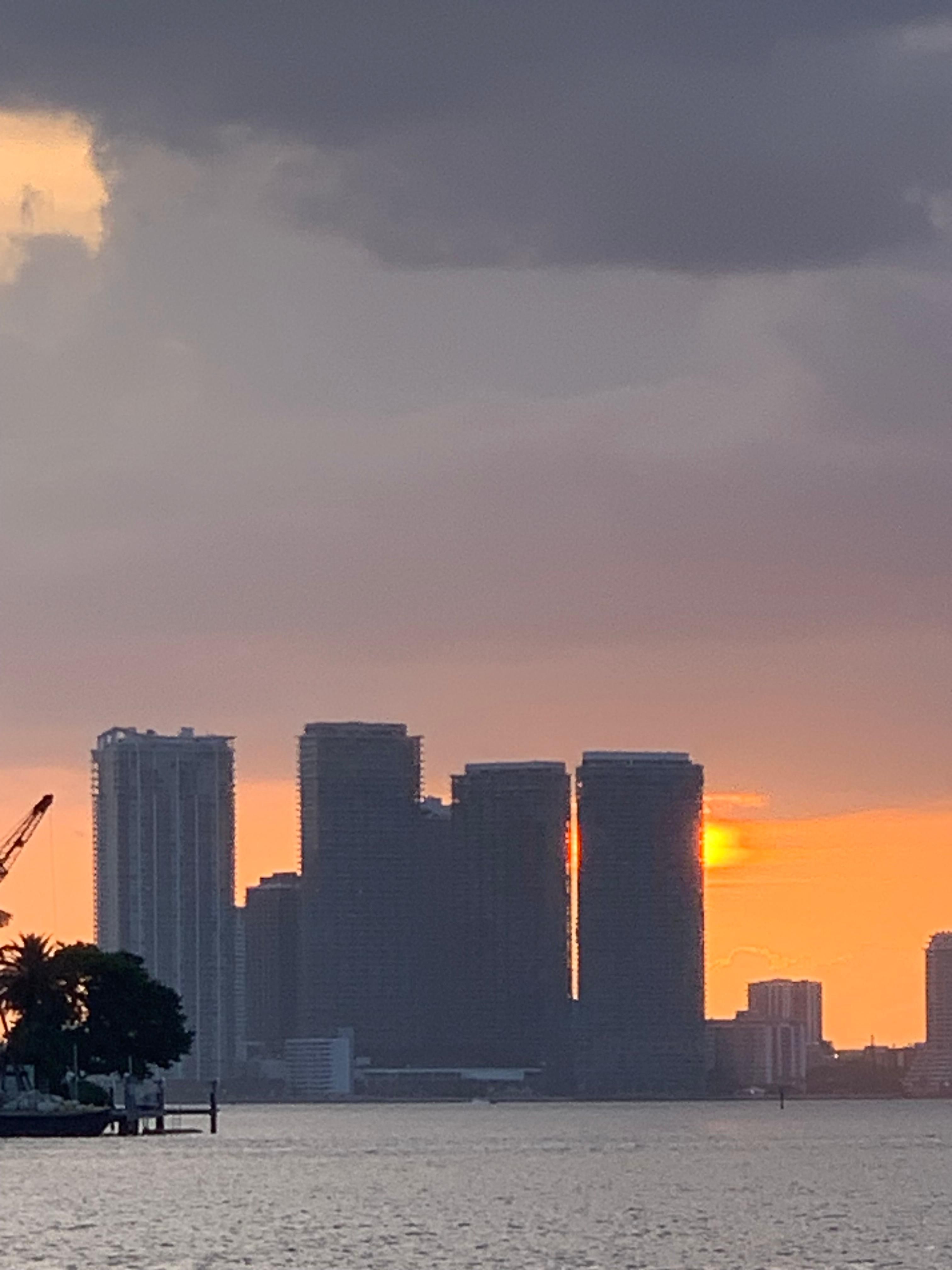 City skyline silhouetted against a vibrant orange sunset over water, with the sun visible between buildings.