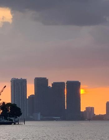 City skyline silhouetted against a vibrant orange sunset over water, with the sun visible between buildings.