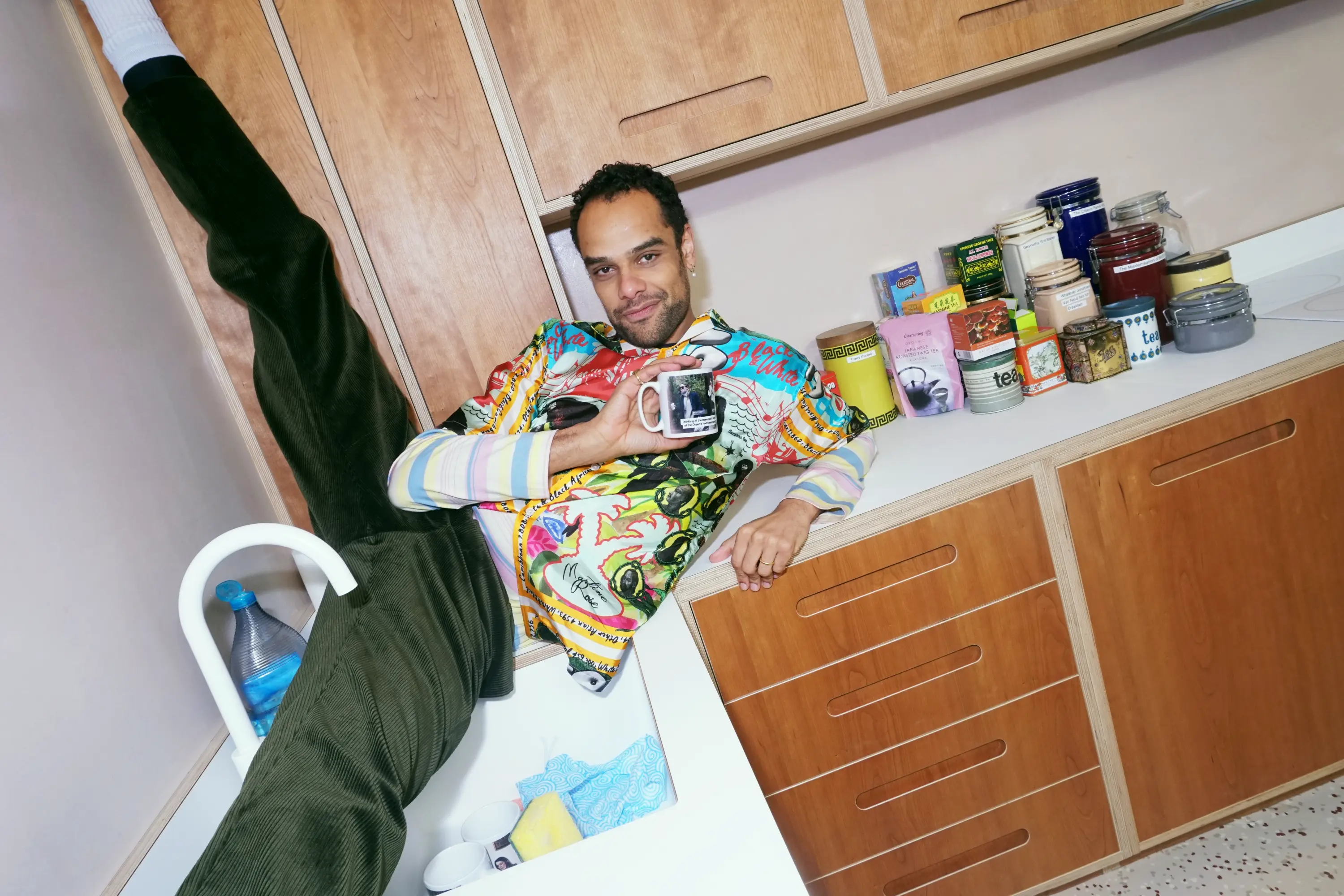 A smiling man in a colorful shirt rests on a kitchen counter, his legs in the sink, holding a mug, with various jars and packages beside him.