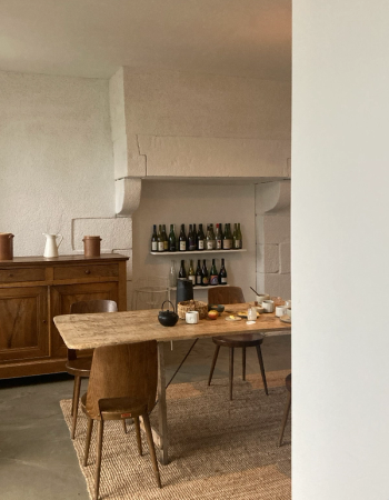 A rustic dining room features a long wooden table and chairs on a woven rug, a stone fireplace with shelves of bottles in the background, and a wooden sideboard to the left. The table is set with a black teapot and other meal items.