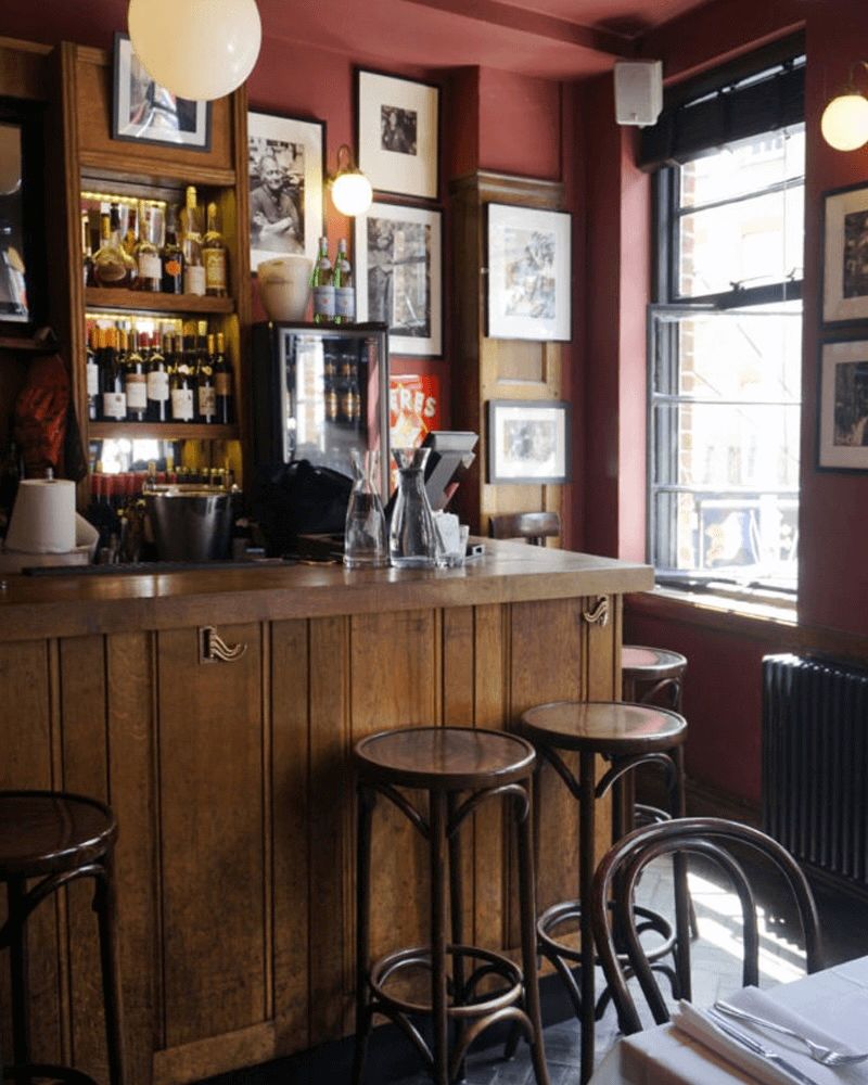 A cozy bar or restaurant interior featuring a wooden counter with stools, stocked shelves, red walls with framed art, and a large window.