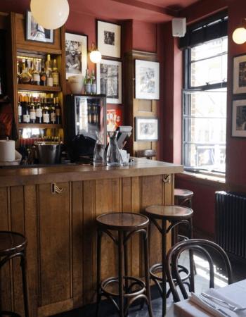 A cozy bar or restaurant interior featuring a wooden counter with stools, stocked shelves, red walls with framed art, and a large window.