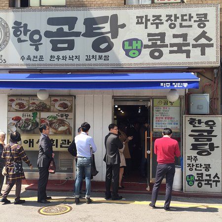 People wait to enter a Korean restaurant with a large sign, a blue awning, and menu pictures on the wall.