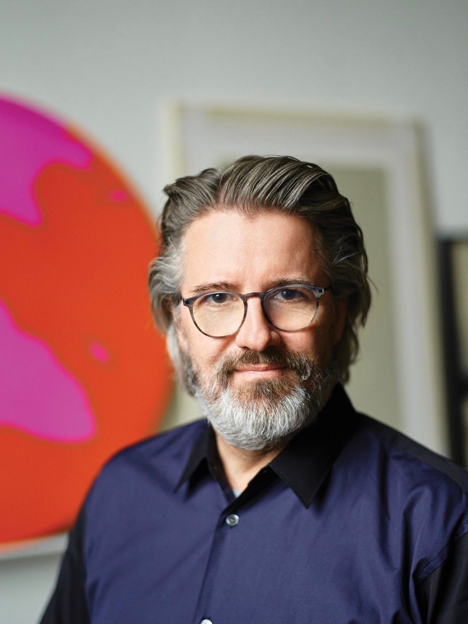 A man with gray hair and beard, wearing glasses and a dark blue shirt, smiles slightly in front of abstract pink and orange art.