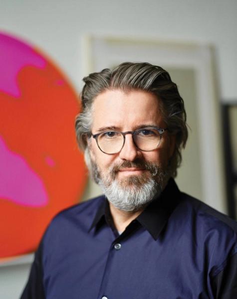A man with gray hair and beard, wearing glasses and a dark blue shirt, smiles slightly in front of abstract pink and orange art.
