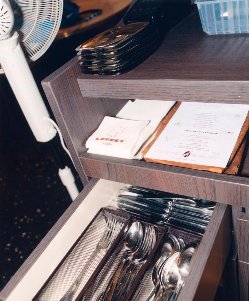 A wooden service cabinet with an open drawer of organized silverware, a shelf holding napkins and a menu, and a white fan nearby.
