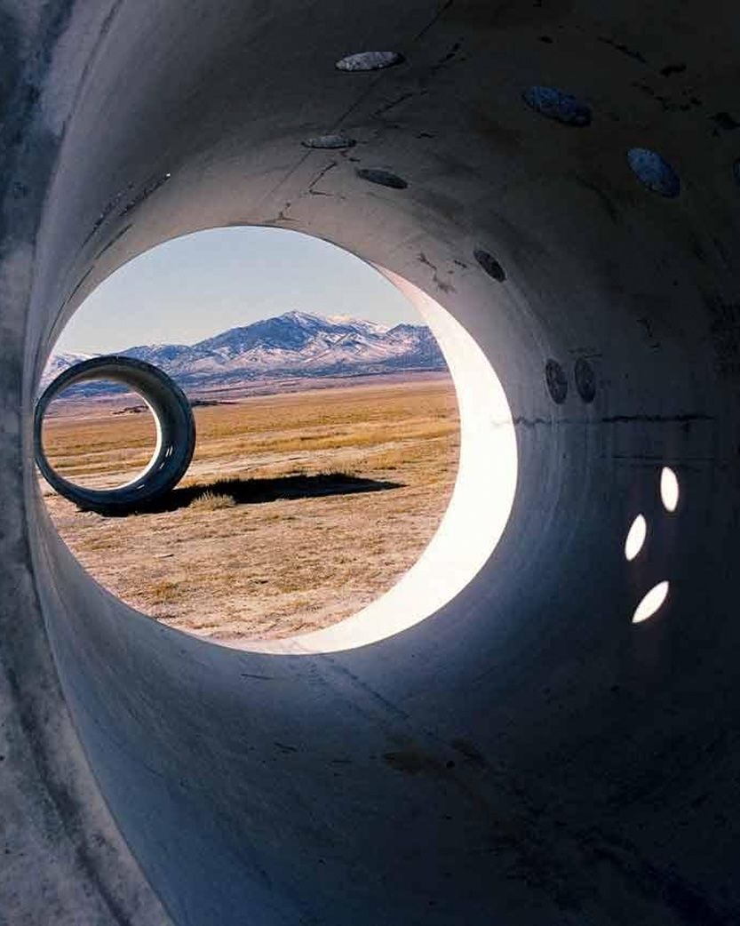 View from inside a large concrete pipe looking out to a desert landscape with a smaller concrete ring and distant mountains.
