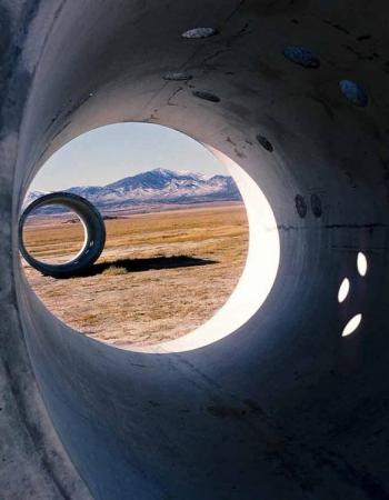 View from inside a large concrete pipe looking out to a desert landscape with a smaller concrete ring and distant mountains.