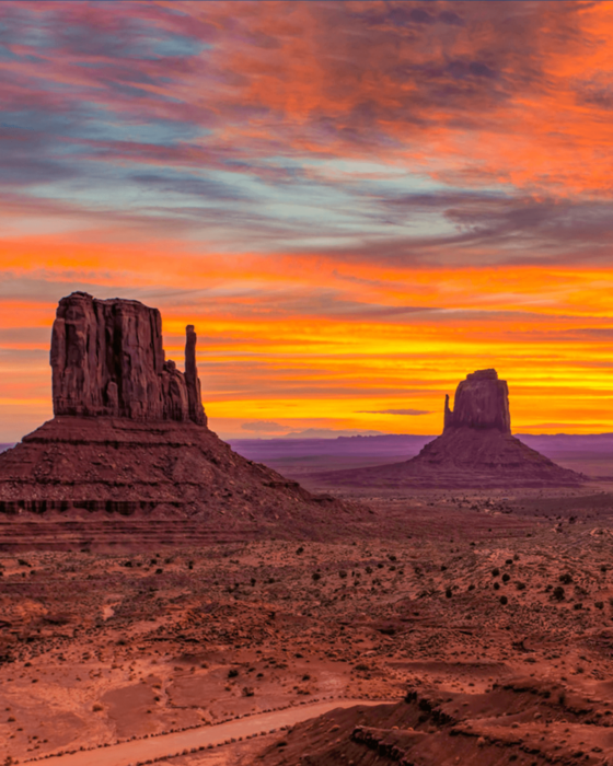 Monument Valley at sunset, with two red rock buttes against a fiery orange and yellow sky.