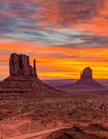 Monument Valley at sunset, with two red rock buttes against a fiery orange and yellow sky.