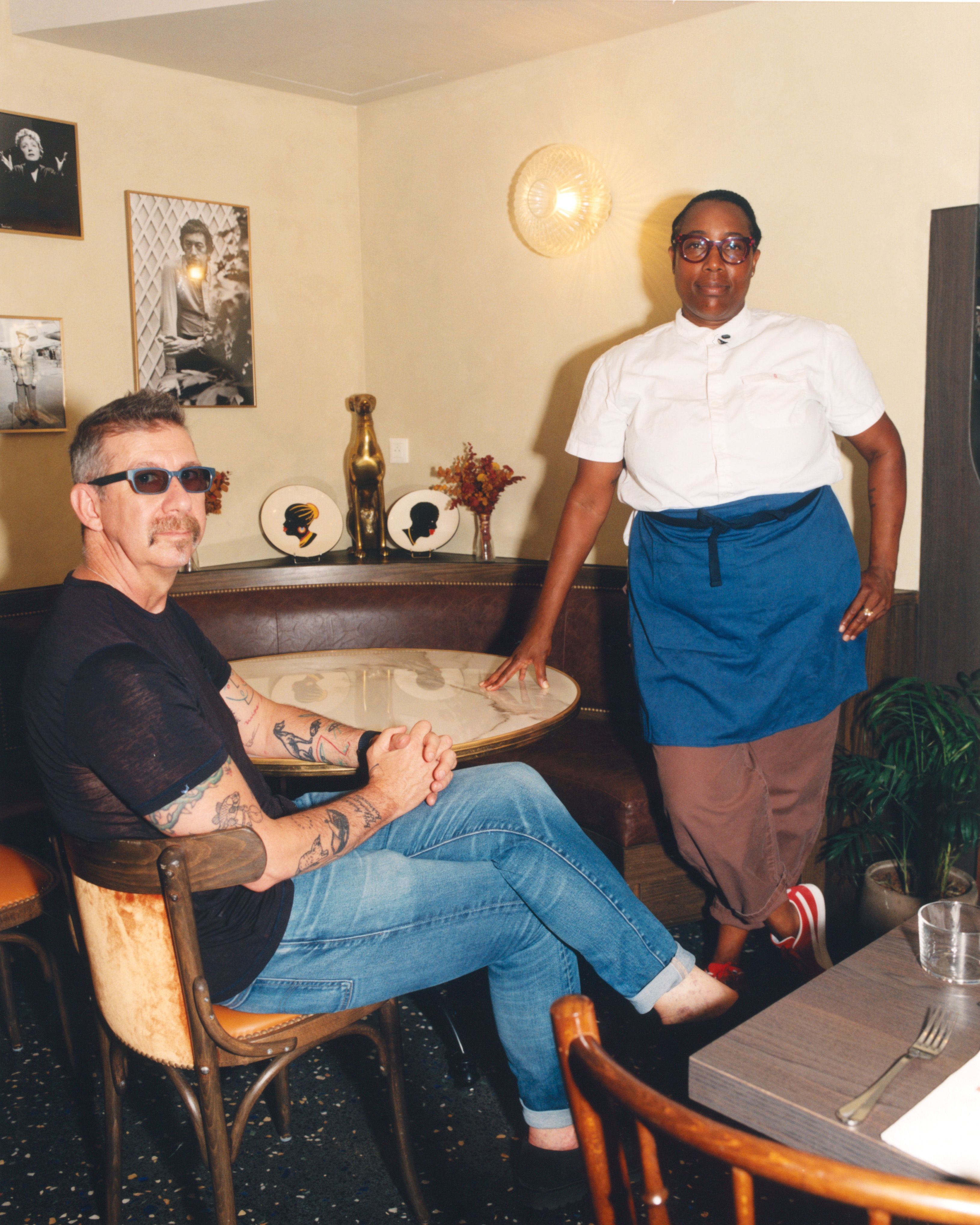 A man in sunglasses sits at a table while a woman in a chef's uniform stands beside him in a decorated room.