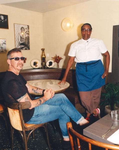 A man in sunglasses sits at a table while a woman in a chef's uniform stands beside him in a decorated room.
