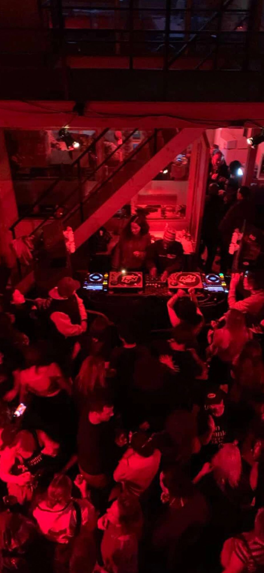 Two DJs perform for a crowd dancing in a club illuminated by red lights.