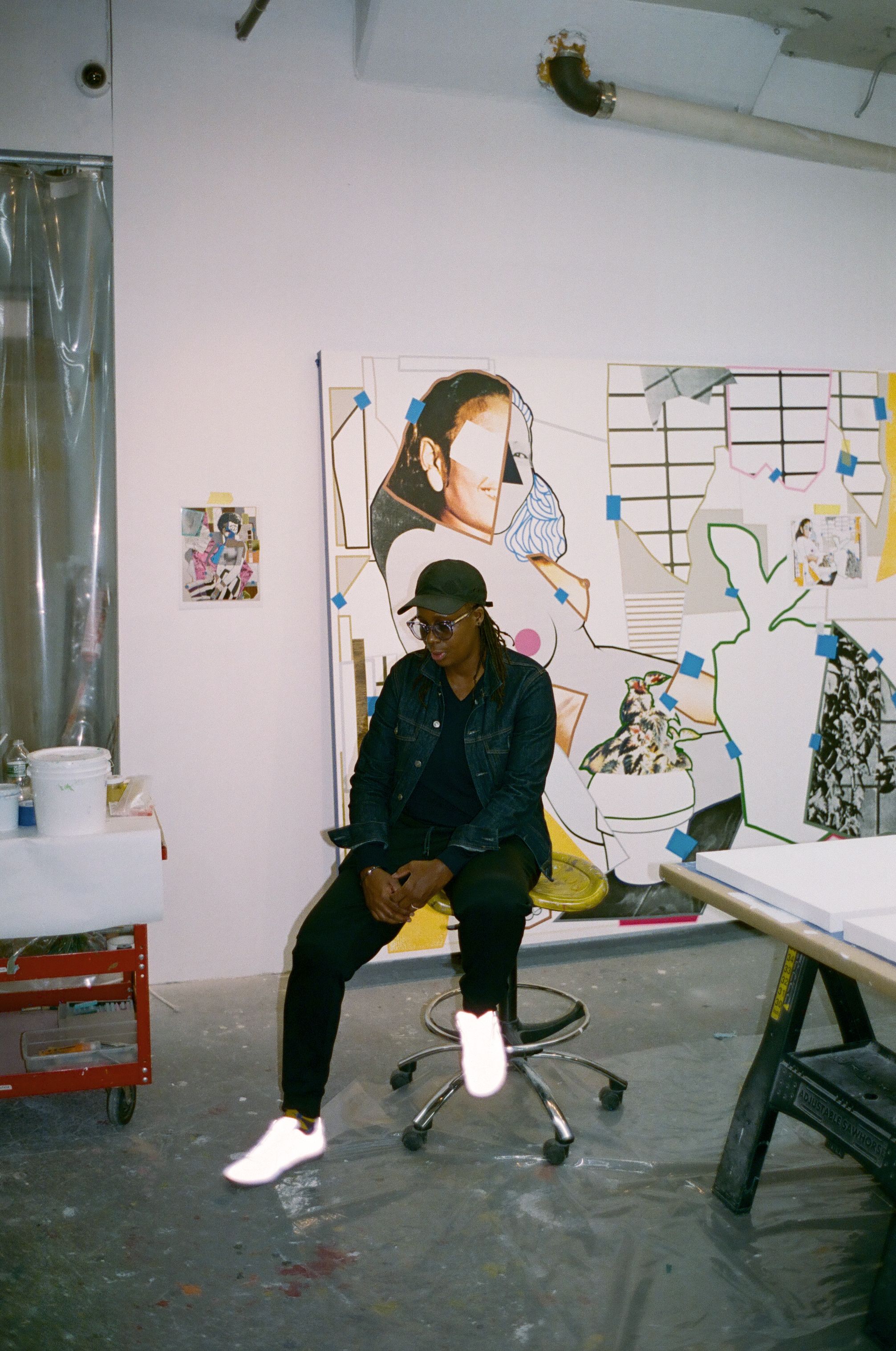 A person in a denim jacket, cap, and reflective white sneakers sits on a stool in an art studio, in front of a large, fragmented collage artwork.