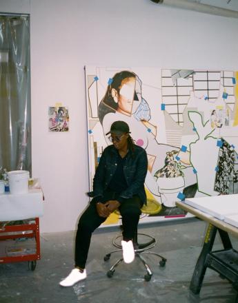 A person in a denim jacket, cap, and reflective white sneakers sits on a stool in an art studio, in front of a large, fragmented collage artwork.