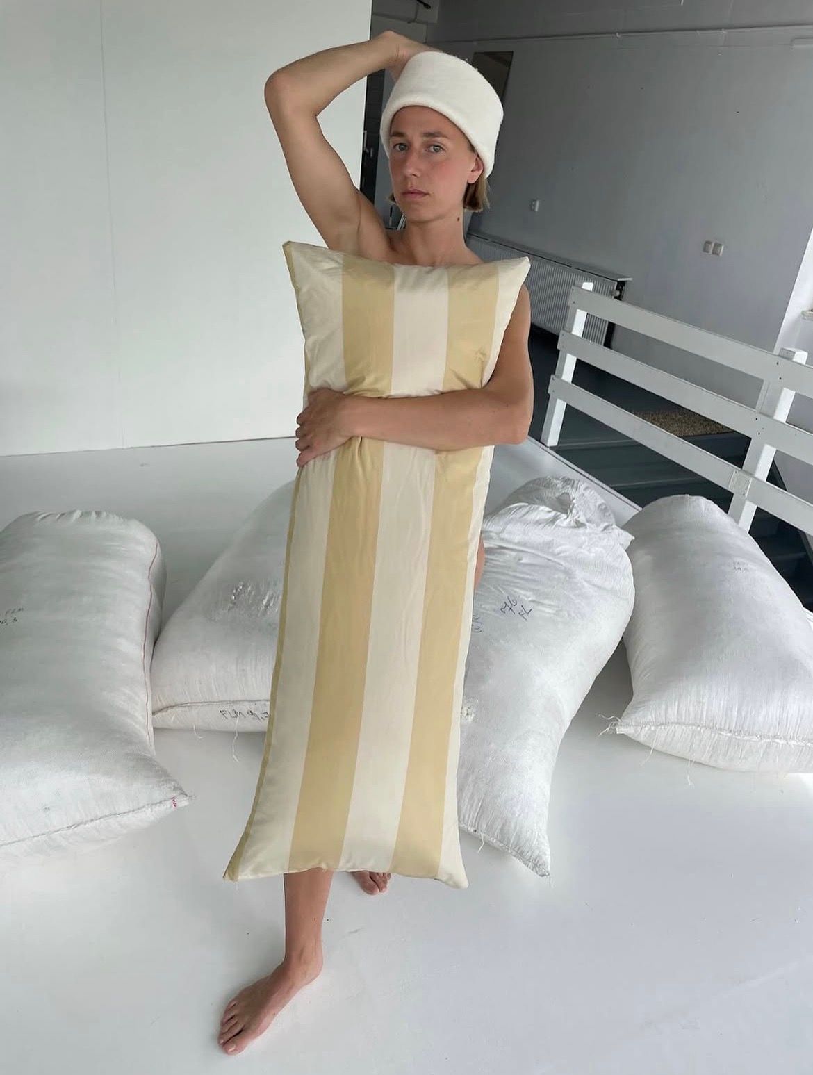 A barefoot person in a white towel hat holds a long yellow and white striped pillow over their body, surrounded by white sacks.