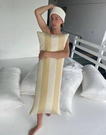 A barefoot person in a white towel hat holds a long yellow and white striped pillow over their body, surrounded by white sacks.