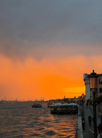 Vibrant orange sunset over a cityscape on a waterfront with boats and people on a promenade.