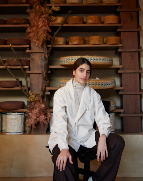 A young woman in a cream jacket sits on a stool in front of wooden shelves filled with wheels of cheese and dried foliage.