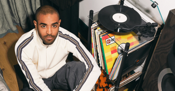 A bearded man in a cream tracksuit sits on the floor, looking at the camera, next to a turntable and a stack of records.