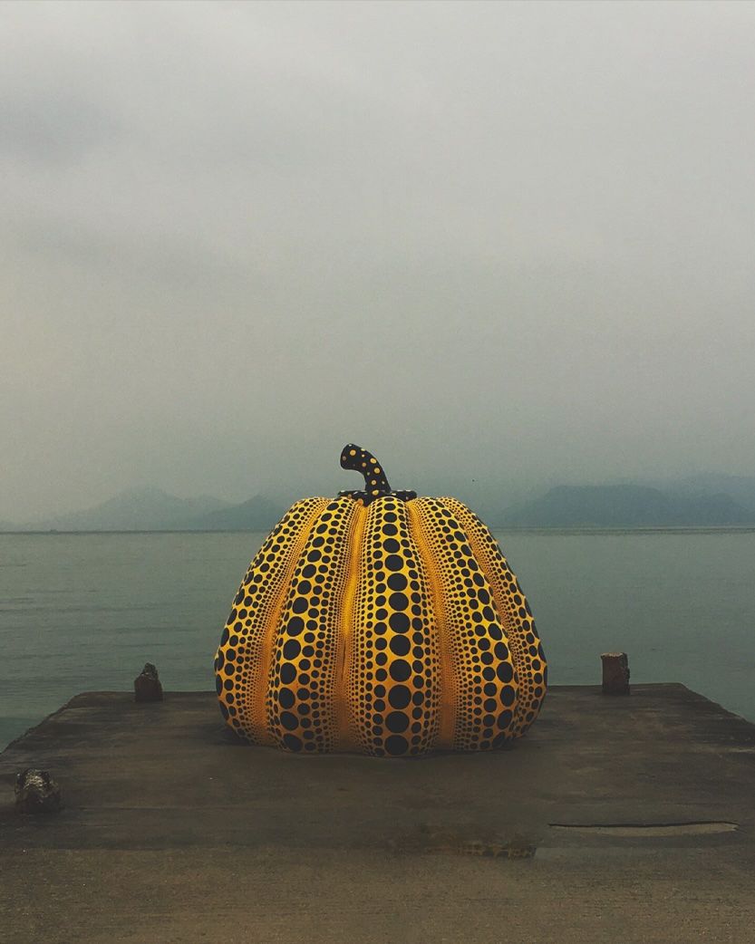 A large yellow pumpkin sculpture with black polka dots sits on a pier by the sea, with distant hazy mountains under a gray sky.