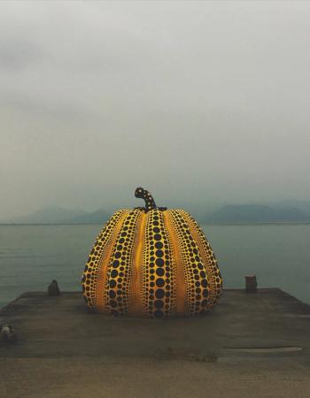 A large yellow pumpkin sculpture with black polka dots sits on a pier by the sea, with distant hazy mountains under a gray sky.