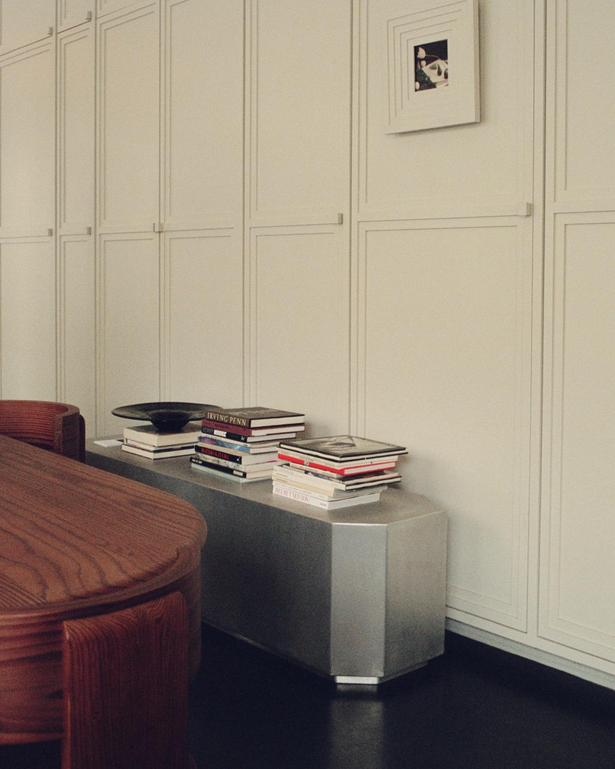 A modern room with a wooden table, a metallic bench with books, and white paneled walls.