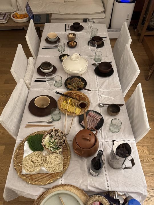 An overhead view of a dining table covered with a white tablecloth, laden with a variety of Korean dishes, raw ingredients for a meal, and individual place settings.