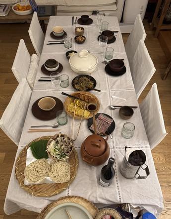 An overhead view of a dining table covered with a white tablecloth, laden with a variety of Korean dishes, raw ingredients for a meal, and individual place settings.