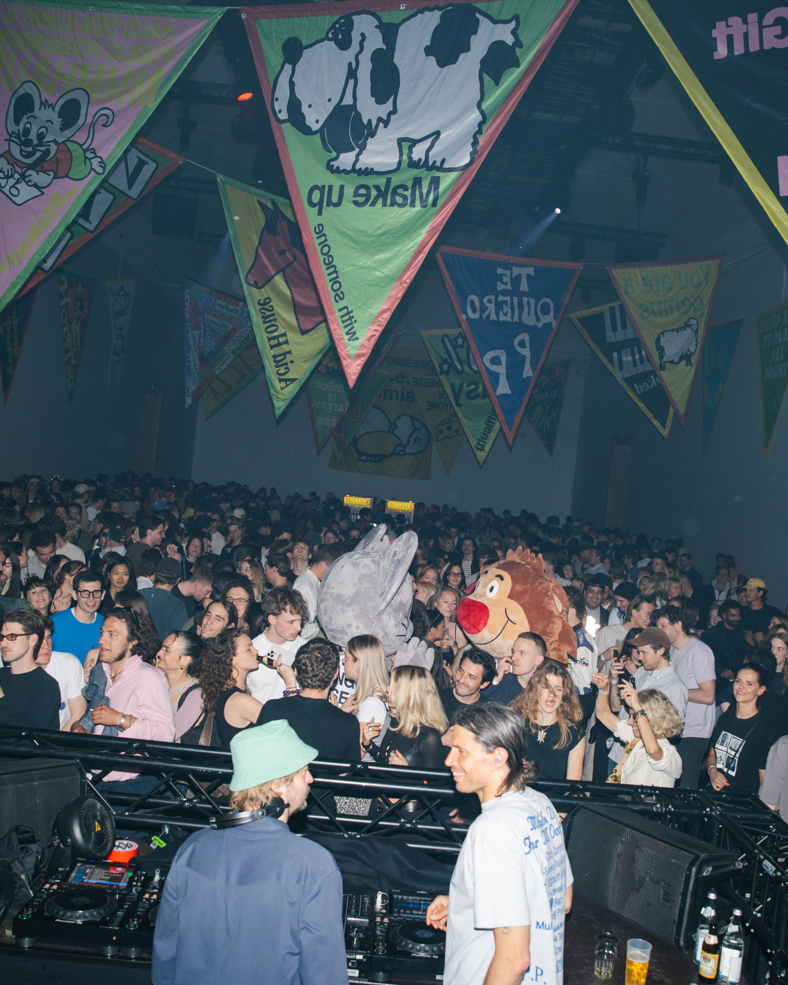 A lively event with a large crowd, two DJs, and colorful banners hanging from the ceiling. Several attendees wear animal costume heads.