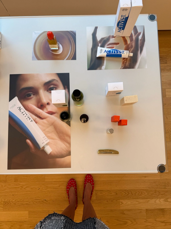 Overhead view of a white table displaying beauty products, including Activist brand tubes and boxes, alongside promotional images, with a person's feet in red studded shoes visible at the bottom.