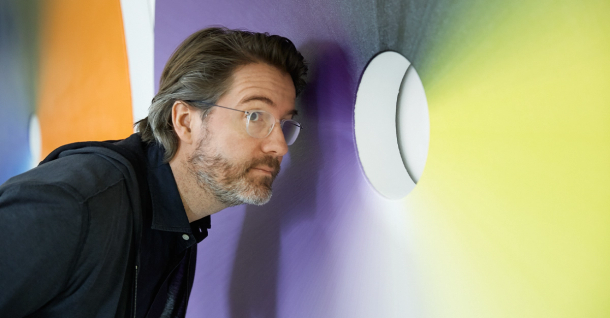 A man with glasses and a beard looks intently into a glowing circular opening in a purple and yellow wall.