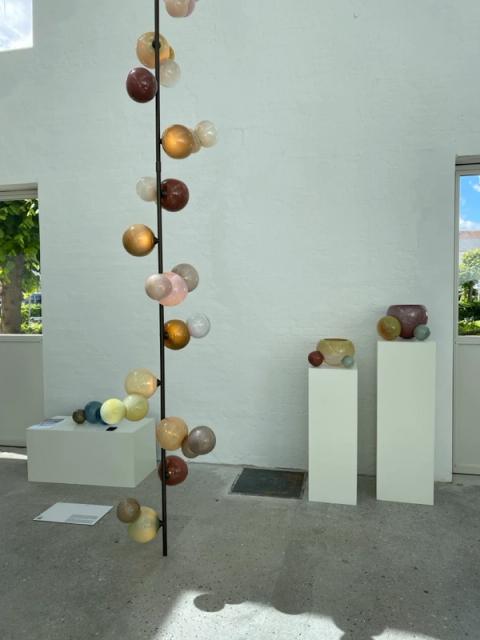 A contemporary art installation in a white gallery room features a tall sculpture of colorful glass spheres on a pole and various glass vessels displayed on white pedestals.