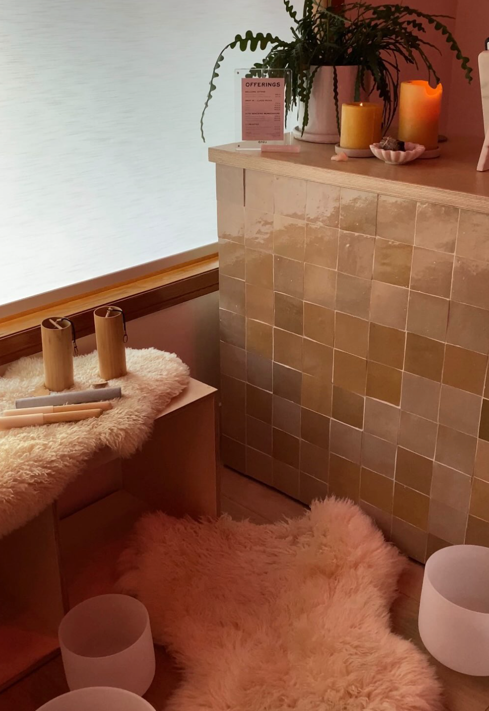 A serene wellness space with a shimmering tiled counter holding candles and a plant. Fluffy rugs on the floor feature wooden sound healing tools and white singing bowls.