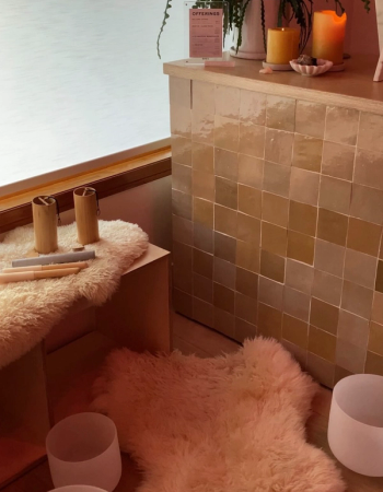A serene wellness space with a shimmering tiled counter holding candles and a plant. Fluffy rugs on the floor feature wooden sound healing tools and white singing bowls.