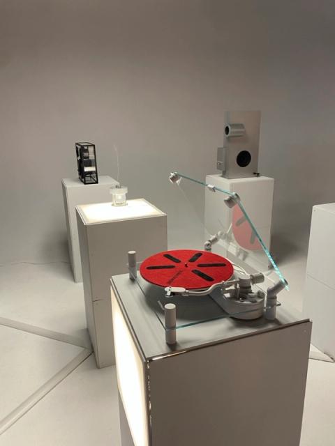 A minimalist art exhibit featuring a modern record player with a red mat, abstract sculptures, and a vessel emitting a wisp, all on white pedestals in a bright white room.