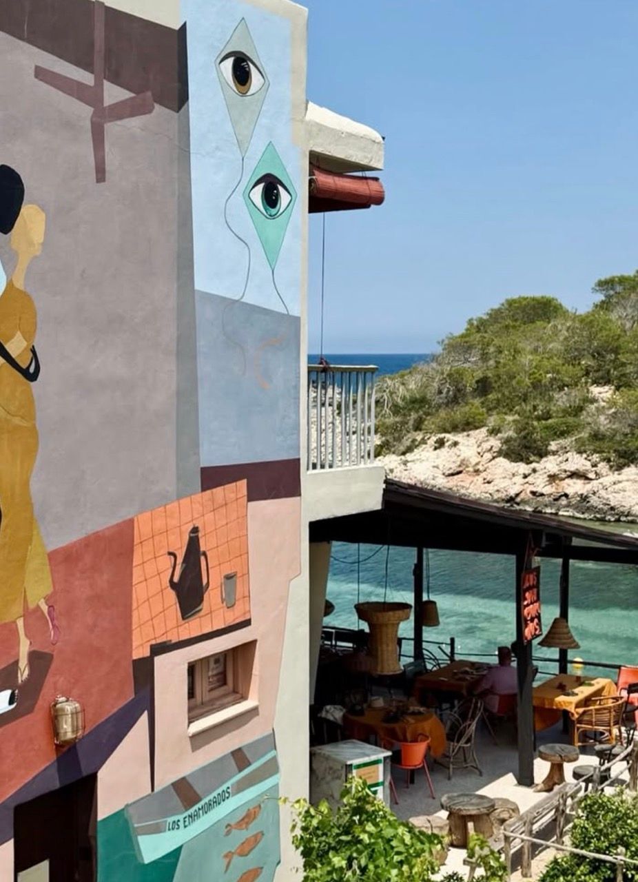 A vibrant building with abstract murals, including eyes and figures, stands next to an outdoor restaurant overlooking a tranquil turquoise sea and a green, rocky coastline.