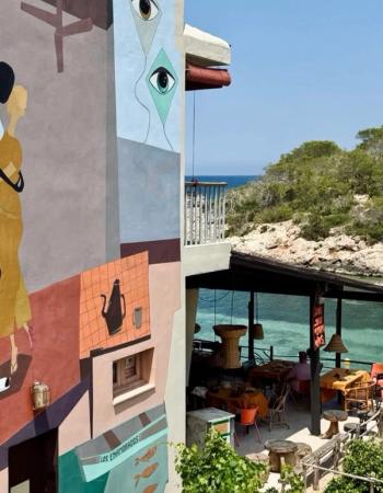 A vibrant building with abstract murals, including eyes and figures, stands next to an outdoor restaurant overlooking a tranquil turquoise sea and a green, rocky coastline.