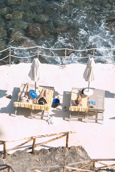 Two people sunbathe on lounge chairs under umbrellas on a concrete patio next to the sea.