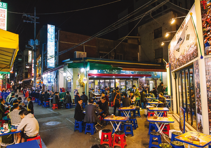 Bustling Korean street at night, with many people dining and drinking at outdoor tables and stools.