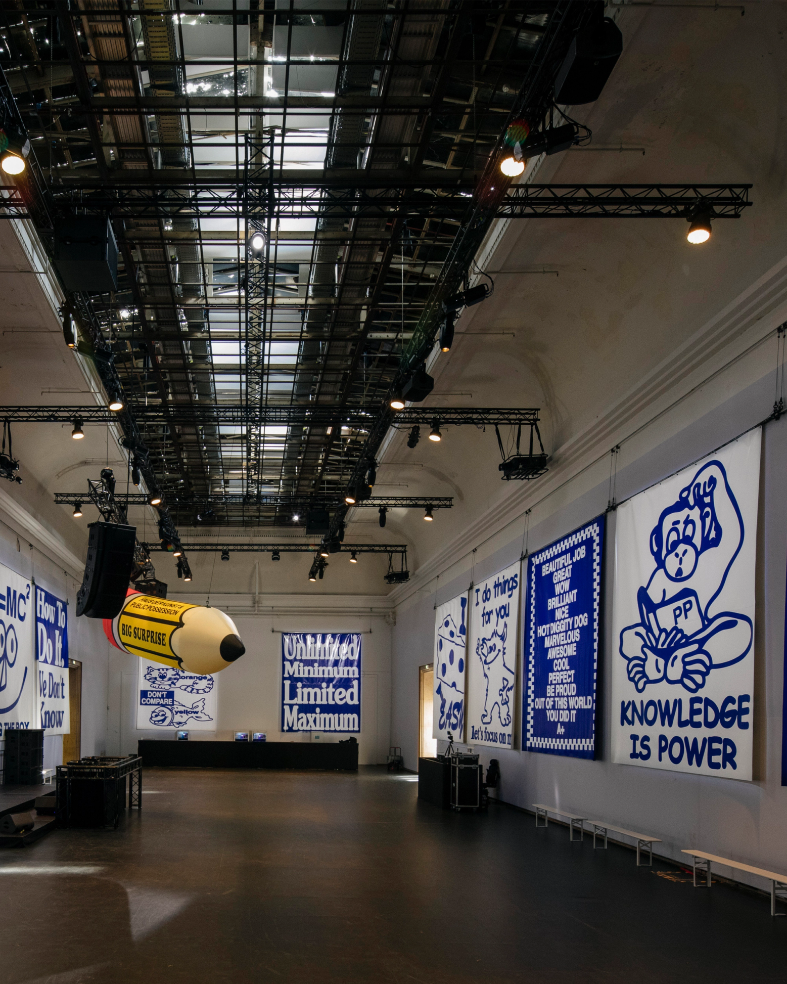 A large, empty hall with a high, grid-trussed ceiling and lights, decorated with white banners featuring blue illustrations and text, and a giant yellow inflatable pencil hanging overhead.
